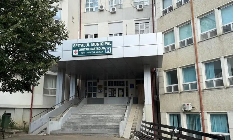 Intrarea unui spital este accesibilă printr-o scară de beton și o rampă, sub un panou alb cu text negru și un simbol medical roșu.