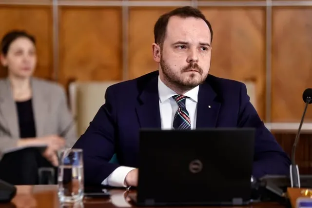 Un bărbat cu barbă și costum închis la culoare stă la o masă cu un laptop în față, în timp ce o femeie stă în fundal.