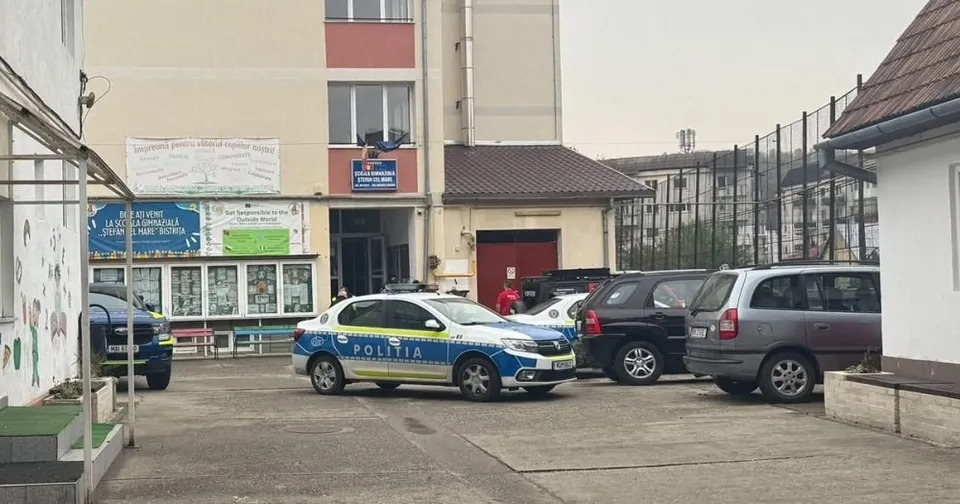 Două mașini de poliție și trei autoturisme sunt parcate în fața unei clădiri cu ferestre și un pridvor.