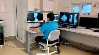 O femeie cu o cămașă albastră stă pe un scaun de birou în fața mai multor monitoare care afișează imagini medicale într-o încăpere cu plăci ceramice.