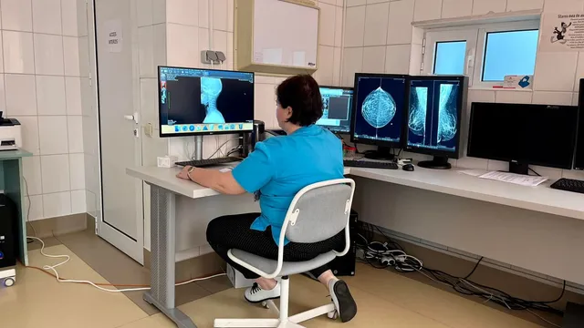 O femeie cu o cămașă albastră stă pe un scaun de birou în fața mai multor monitoare care afișează imagini medicale într-o încăpere cu plăci ceramice.