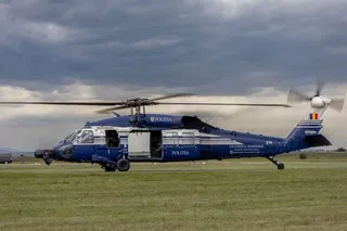 Un elicopter albastru cu inscripția Politia stă aterizat pe o câmpie de iarbă sub un cer înnorat.