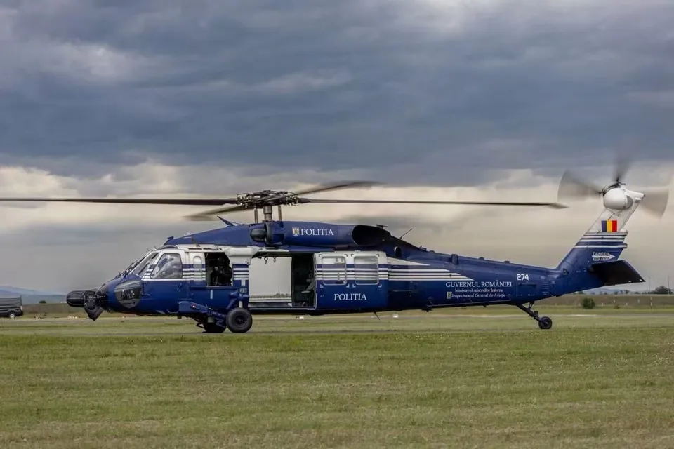 Un elicopter albastru cu inscripția Politia stă aterizat pe o câmpie de iarbă sub un cer înnorat.