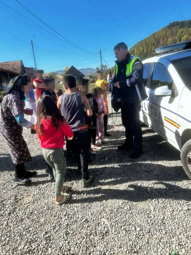 Un polițist în uniformă cu vestă reflectorizantă stă lângă o mașină albă, în timp ce un grup de copii stă pe un teren cu pietriș.