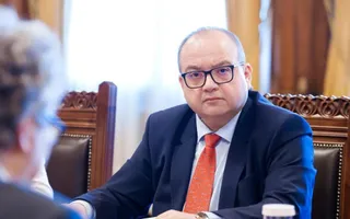 Un bărbat cu ochelari și cravată roșie stă la o masă de lemn lângă un scaun cu spătar înalt.