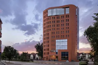 O clădire înaltă, de culoare portocalie, cu ferestre rectangulare și o secțiune frontală cu panouri de sticlă, sub un cer cu nori la apus.