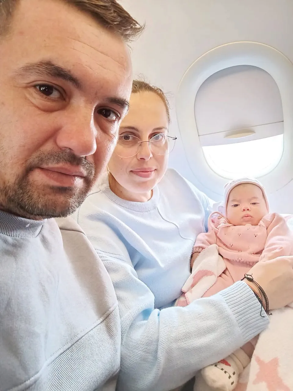 Un bărbat, o femeie cu ochelari și un bebeluș îmbrăcat în roz sunt așezați lângă o fereastră de avion.