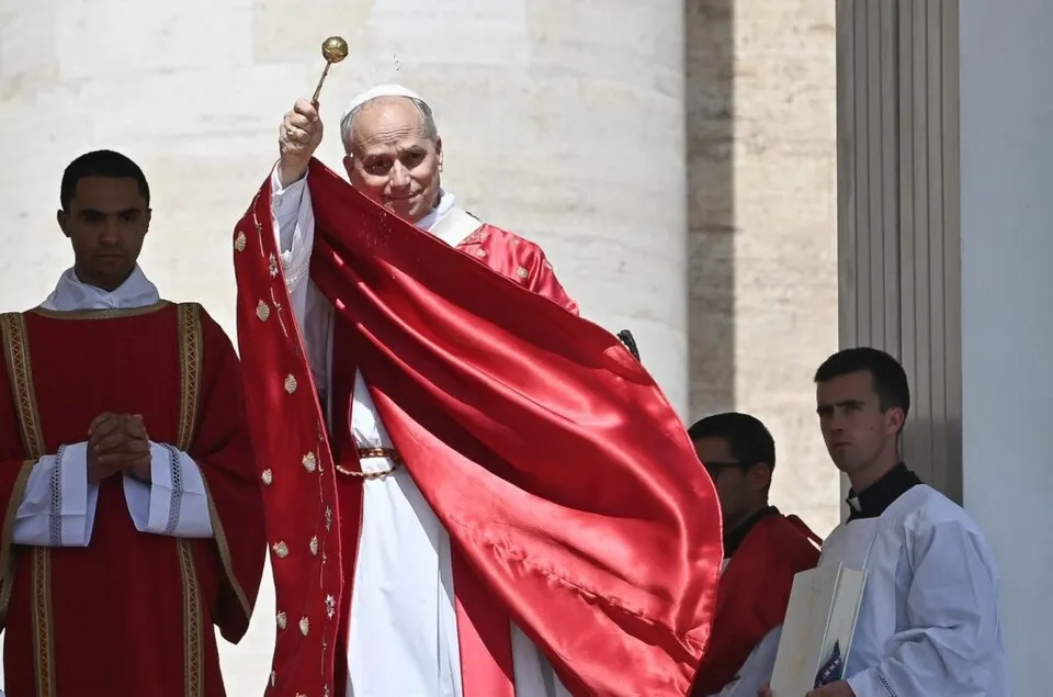 Un bărbat în haine albe și roșii ridică un obiect auriat, fiind înconjurat de alți doi bărbați îmbrăcați în haine liturgice.