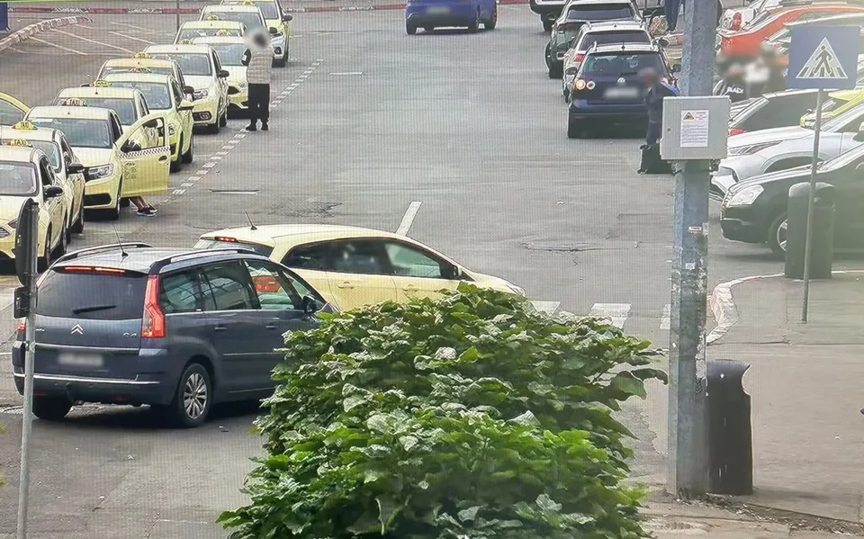 Mai multe mașini sunt parcate pe o stradă lângă un gard și un bușten, în timp ce un vehicul alb circulă pe carosabil.