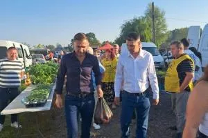 Câțiva bărbați merg pe un teren verde lângă mese cu legume, în timp ce alți oameni stau în fundal sub cerul senin.