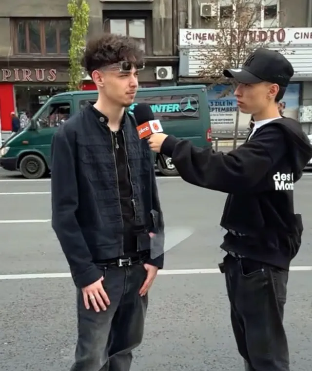 Un bărbat cu ochelari de soare și o jachetă neagră este intervievat de un alt bărbat cu șapcă și hanorac negru pe un drum asfaltat.