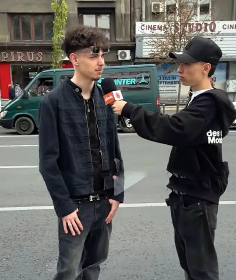 Un bărbat cu ochelari de soare și o jachetă neagră este intervievat de un alt bărbat cu șapcă și hanorac negru pe un drum asfaltat.