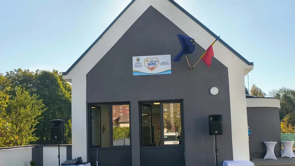 O clădire gri cu acoperiș în formă de triunghi prezintă un panou informativ și două steaguri pe fațadă, lângă două boxe de sunet.