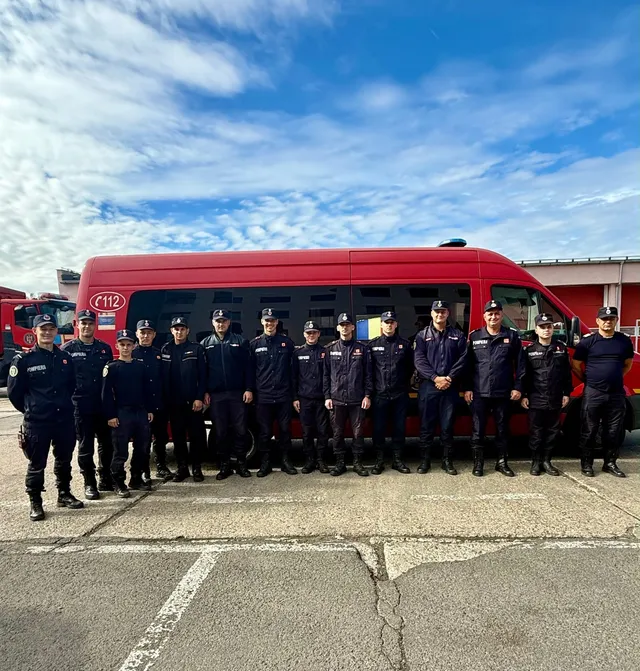 Un grup de oameni în uniforme închise stau în linie în fața unui vehicul de serviciu de culoare roșie sub un cer cu nori.