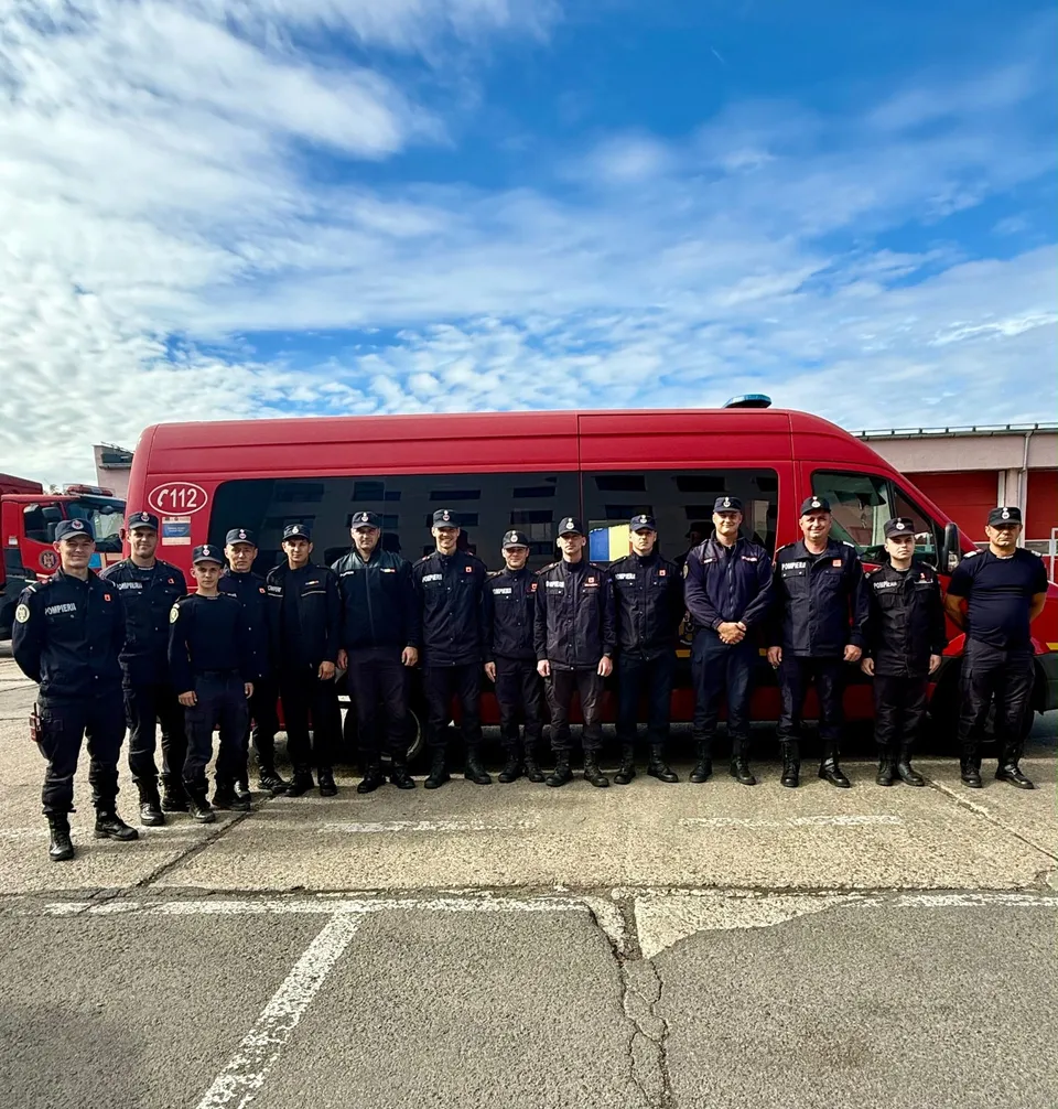Un grup de oameni în uniforme închise stau în linie în fața unui vehicul de serviciu de culoare roșie sub un cer cu nori.