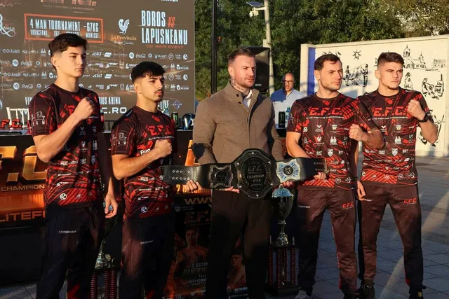 Cinci bărbați stau în linie, patru purtând tricouri negre cu imprimeuri roșii și un bărbat în mijloc ținând un centură de campion.