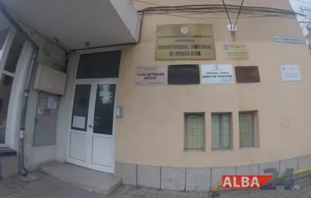 O clădire cu o intrare dotată cu o ușă albă și mai multe plăcuțe informative afișate pe fațada de culoare gălbuie.