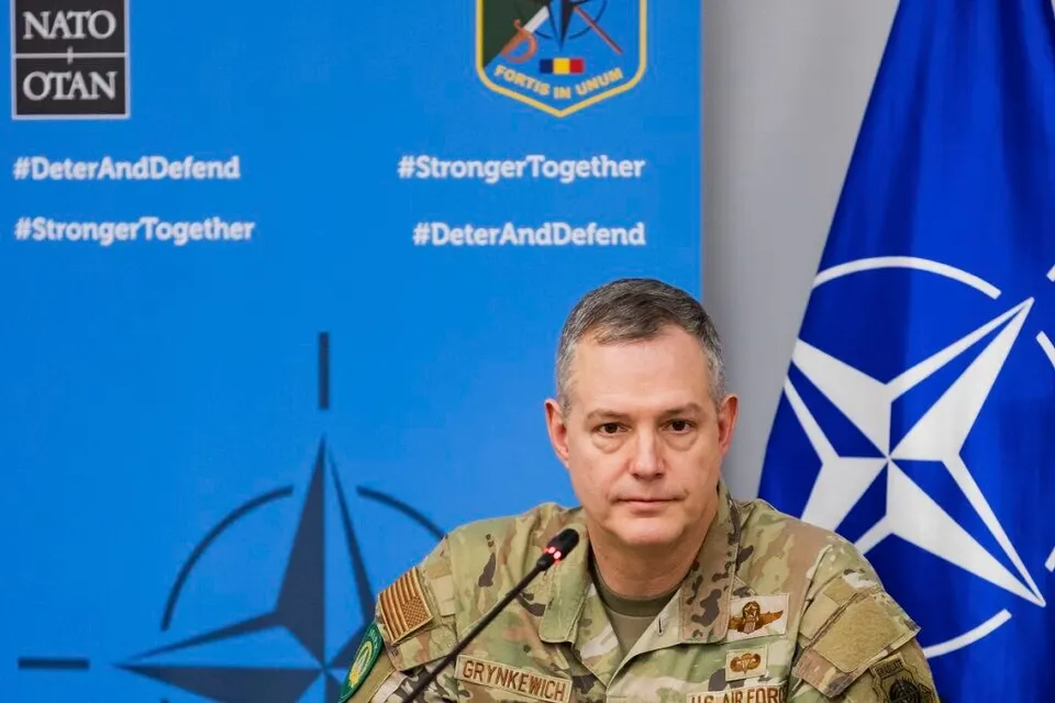 Un bărbat în uniformă militară de tip camuflaj stă în fața unui panou albastru cu logoul NATO și al unui steag albastru.