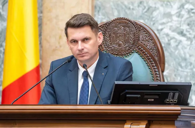 Un bărbat cu părul scurt, purtând sacou albastru, cămașă albă și cravată albastră, stă în spatele unui pupitru de lemn lângă un steag galben, roșu și albastru.