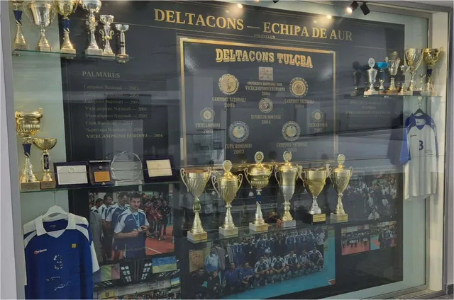 O vitrină neagră expune diverse trofee de argint și aur, un poster cu jucători și un tricou alb cu numărul trei.