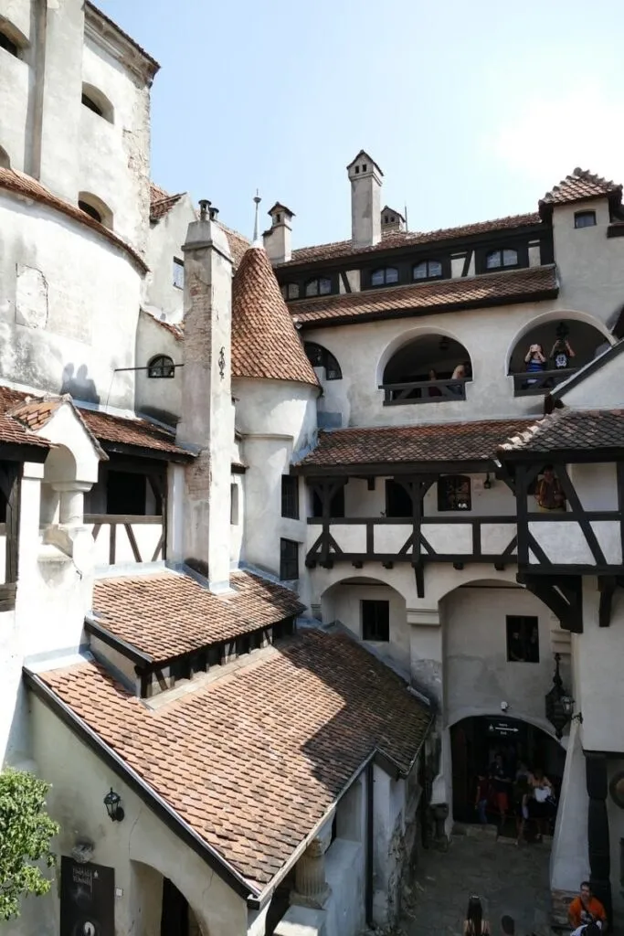 O clădire veche cu ziduri albe, acoperișuri din țiglă brună și ferestre cu arcade înconjoară o curte interioară sub un cer senin.