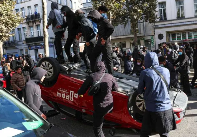 Mai mulți oameni îmbrăcați în haine închise la culoare stau pe un autoturism roșu răsturnat pe o stradă urbană.