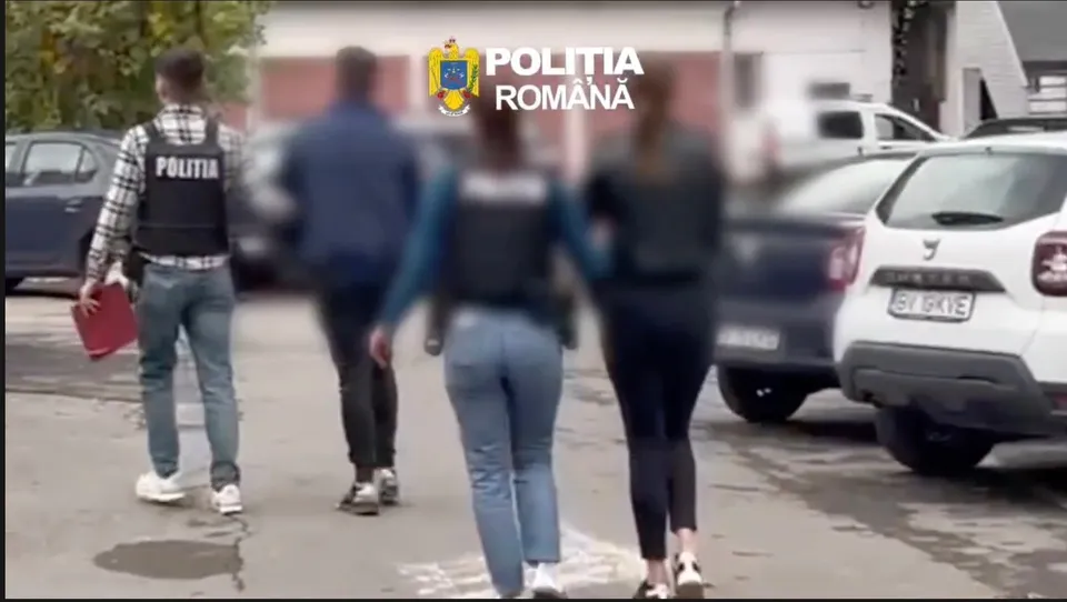 Patru persoane merg pe un drum asfaltat lângă mașini parcate, dintre care una poartă un vest negru cu inscripția POLITIA.
