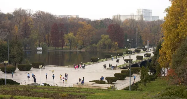 O vedere de la înălțime asupra unui lac înconjurat de copaci cu frunziș colorat, lângă o esplanadă pavată cu persoane care se plimbă.