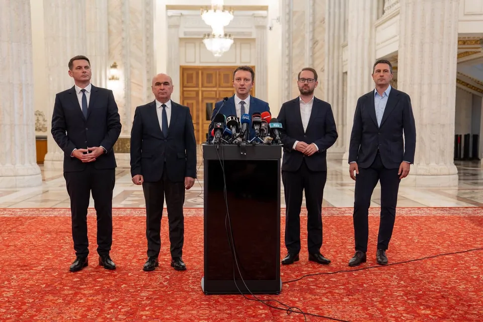 Cinci bărbați în costume stau într-o sală cu coloane, unul la pupitru cu microfoane, pe covor roșu.