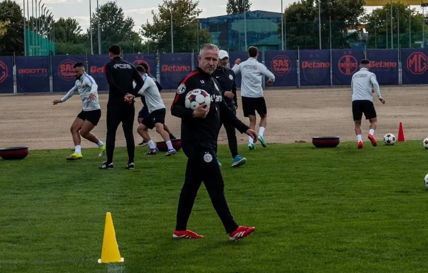 Un bărbat în echipament sportiv negru merge pe un teren de fotbal purtând un balon, în timp ce alți jucători se antrenează în fundal.