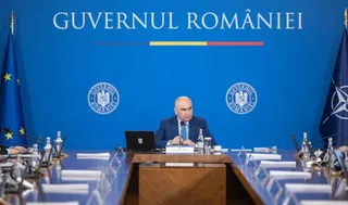 Un bărbat cu costum și cravată stă la o masă de conferințe în fața unui fundal albastru pe care scrie text alb și sunt două embleme circulare.