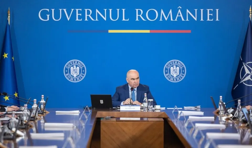 Un bărbat cu costum și cravată stă la o masă de conferințe în fața unui fundal albastru pe care scrie text alb și sunt două embleme circulare.