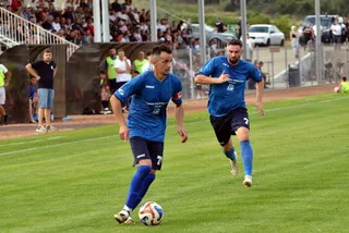 Doi jucători de fotbal în echipamente albastre aleargă pe un teren de gazon verde lângă un balon.