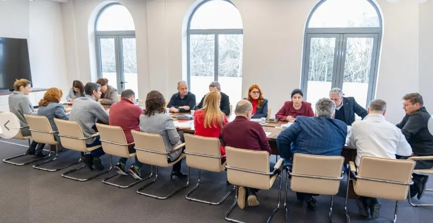 Un grup de persoane este așezat la o masă lungă într-o încăpere cu ferestre mari arcuite, cu spatele la cameră.