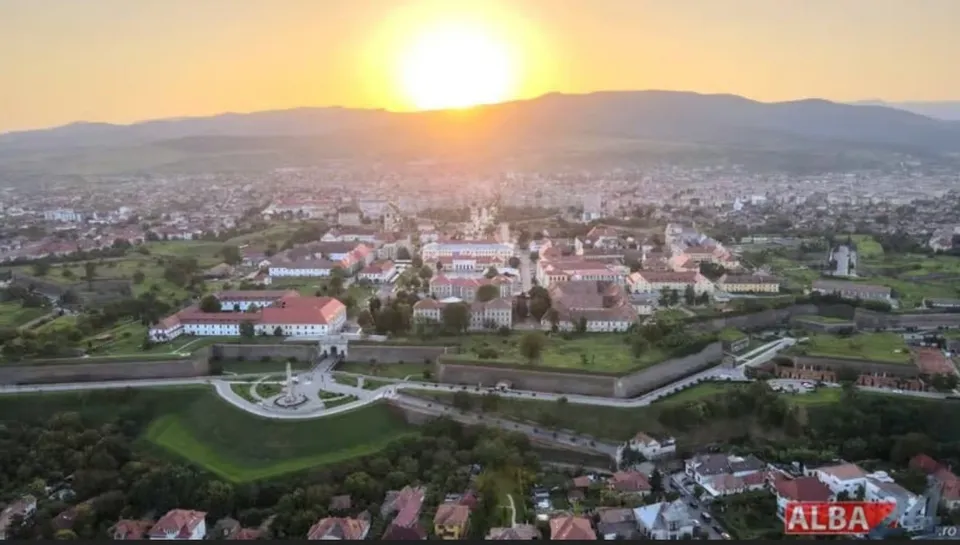 O vedere aeriană a unui oraș cu clădiri și fortificații sub un cer luminos la apus.