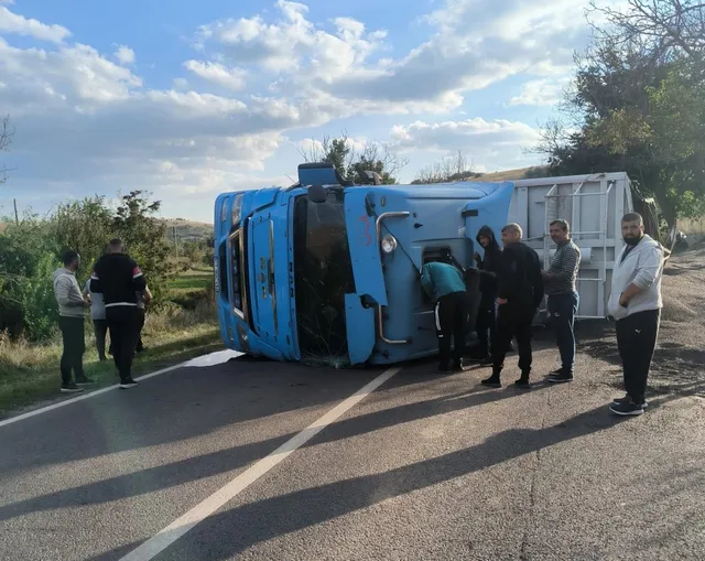 Un camion albastru este răsturnat pe o parte pe marginea unui drum asfaltat, în timp ce mai mulți bărbați stau în apropiere.
