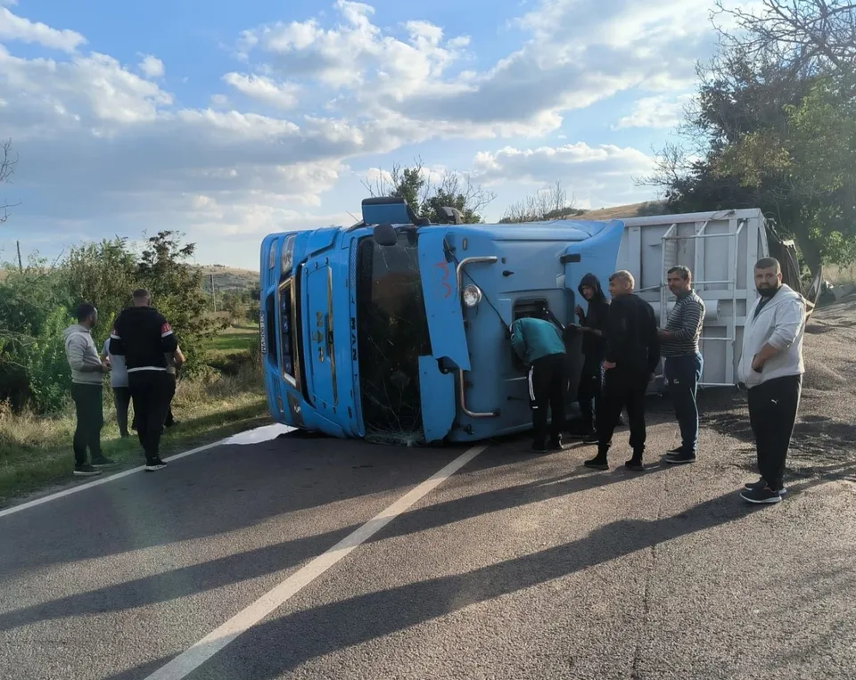 Un camion albastru este răsturnat pe o parte pe marginea unui drum asfaltat, în timp ce mai mulți bărbați stau în apropiere.