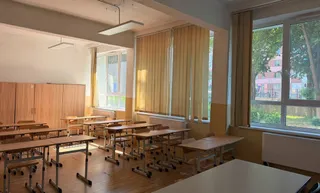O sală de clasă cu bănci de lemn dispuse pe un podea de parchet și ferestre mari cu jaluzele bej.