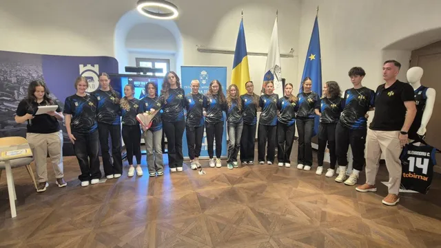 Un grup de femei îmbrăcate în echipamente sportive de culoare închisă stau în rând într-o încăpere cu podea de lemn și steaguri în fundal.