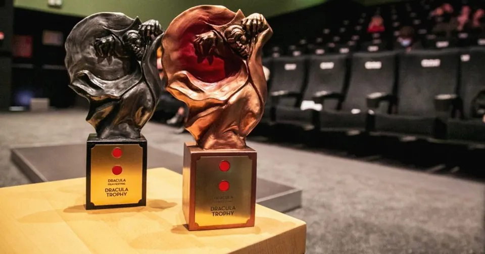 Două trofee sculptate în formă de figuri antropomorfe, una neagră și una bronzată, sunt așezate pe o suprafață lemnoasă în fața unor scaune de cinema.