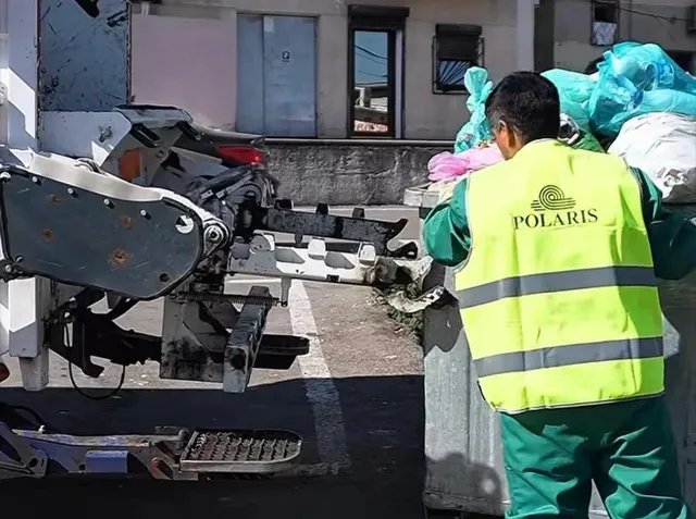 Un bărbat cu vestă reflectorizantă galbenă și pantaloni verzi stă lângă un vehicul utilitar și containere cu saci de gunoi colorați.