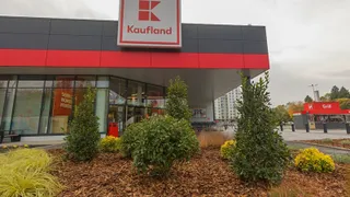 O clădire de magazin cu un panou roșu și alb pe care scrie Kaufland, aflată în spatele unor tufișuri verzi și galbene.