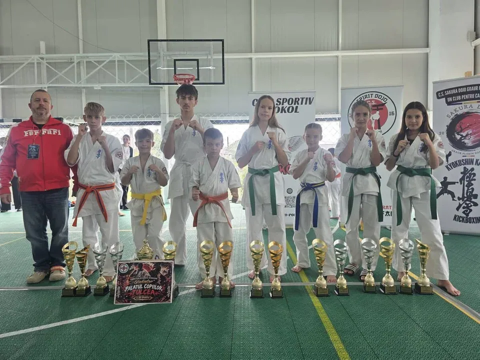 Un grup de copii în echipamente albe de karate și un adult în hanorac roșu stau în spatele unui rând de trofee pe un teren de sport.