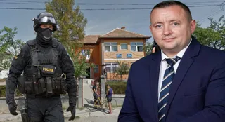 Un bărbat într-un costum albastru stă în prim-plan, în timp ce un polițist echipat cu cască și vestă tactică stă în fundal lângă o clădire portocalie.