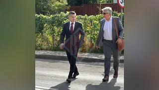 Un bărbat în costum închis și cravată roșie merge pe un trotuar lângă un alt bărbat cu ochelari de soare și sac de piele maro.