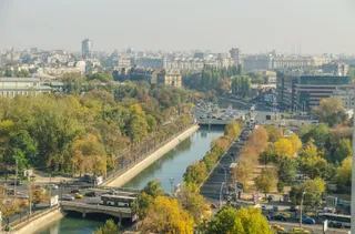 O vedere panoramică asupra unui oraș cu râul Dâmbovița curgând printre arbori cu frunziș galben și verde, lângă drumuri cu mașini circulante.