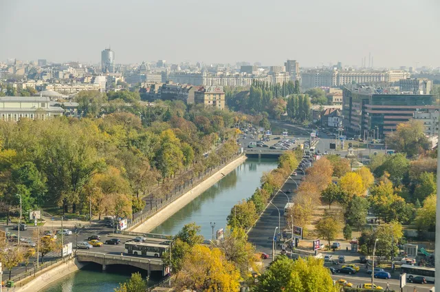 O vedere panoramică asupra unui oraș cu râul Dâmbovița curgând printre arbori cu frunziș galben și verde, lângă drumuri cu mașini circulante.