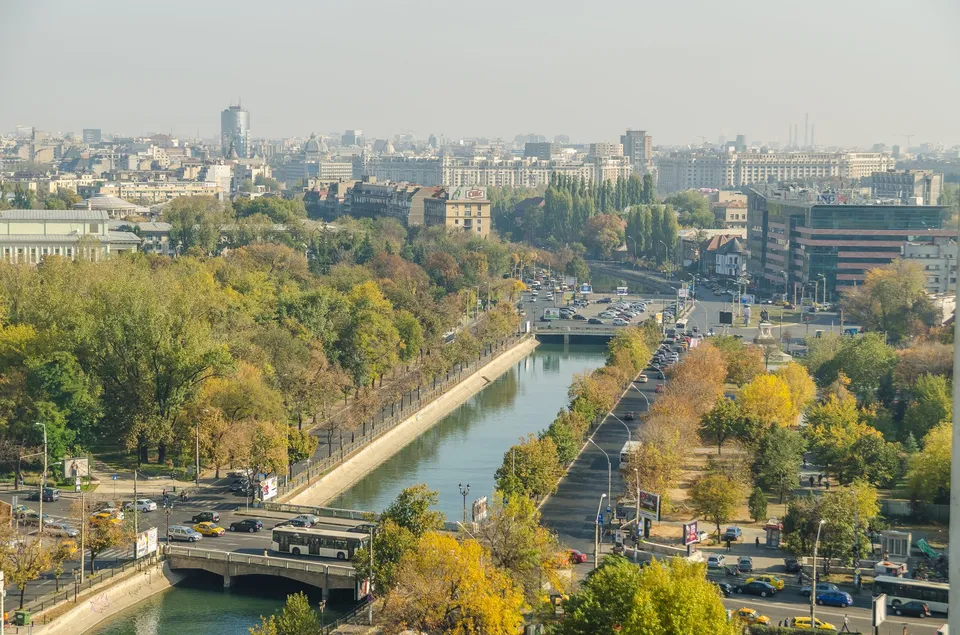 O vedere panoramică asupra unui oraș cu râul Dâmbovița curgând printre arbori cu frunziș galben și verde, lângă drumuri cu mașini circulante.