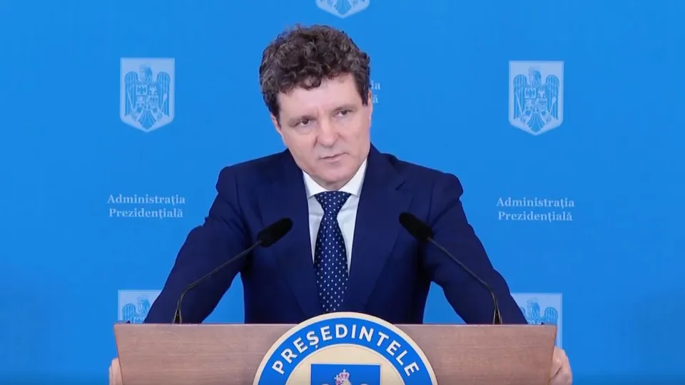 Un bărbat cu părul creț, îmbrăcat în costum închis și cravată cu puncte albe, stă în spatele unui podium în fața unui fundal albastru.