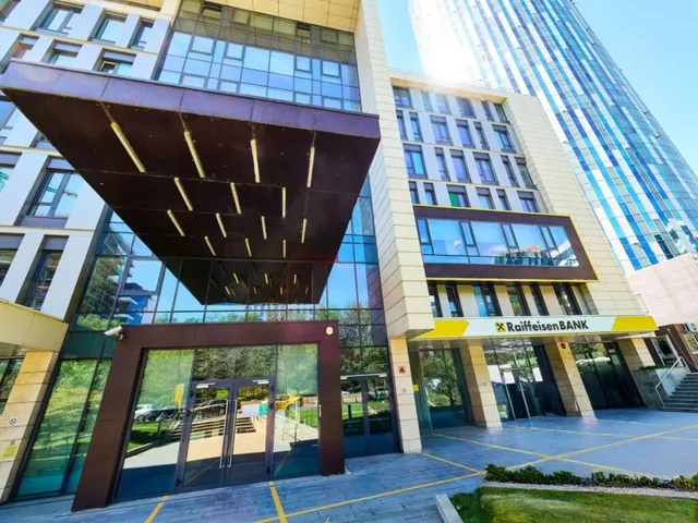 O clădire modernă cu fațadă de sticlă și panouri de piatră prezintă o intrare cu un acoperiș în consolă și un panou galben cu text.
